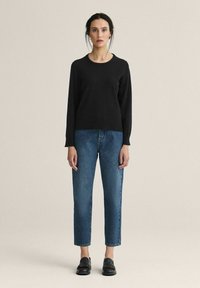 Nainen seisoo yksinkertaisen taustan edessä, yllään musta pitkähihainen neulepaita, siniset farkut ja mustat loafersit.