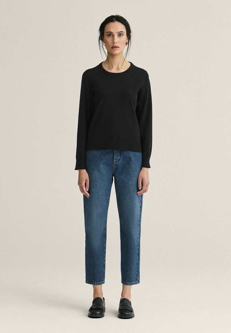 Nainen seisoo yksinkertaisen taustan edessä, yllään musta pitkähihainen neulepaita, siniset farkut ja mustat loafersit.