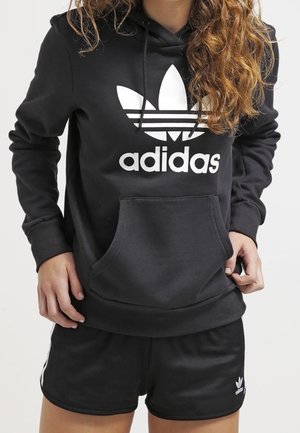 Sort Adidas-hoodie med hvidt trefoil-logo og matchende sorte Adidas-shorts med hvide striber, båret af en person med krøllet hår.