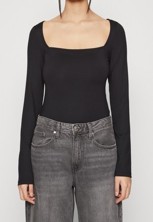 Femme portant un haut noir moulant à manches longues avec un décolleté carré et un jean gris délavé taille haute.