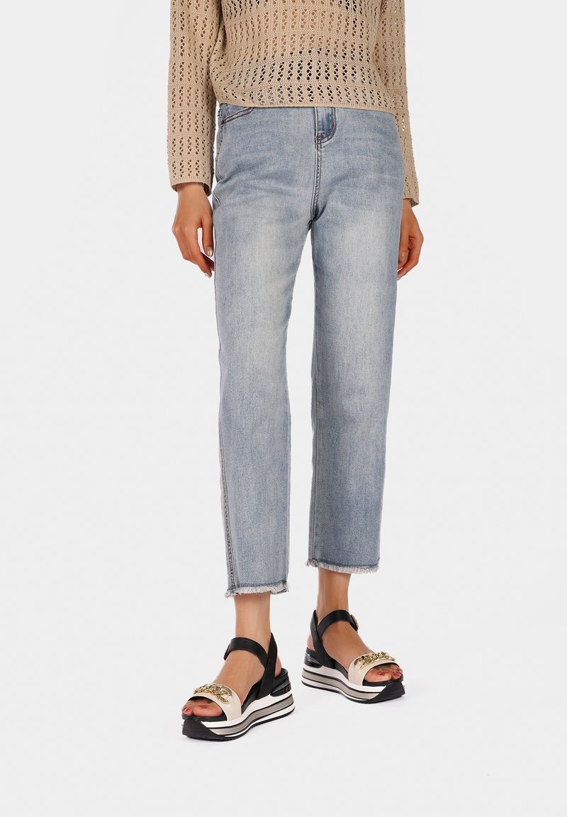 Jeans in denim blu chiaro con orli sfilacciati abbinati a sandali con plateau neri e crema, caratterizzati da un accento decorativo dorato.