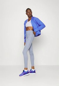 Atlétikus nő áll világosszürke leggingsben, fehér sportmelltartóban, élénk kék dzsekiben és lila futócipőben, oldalt nézve egy sima háttér előtt.