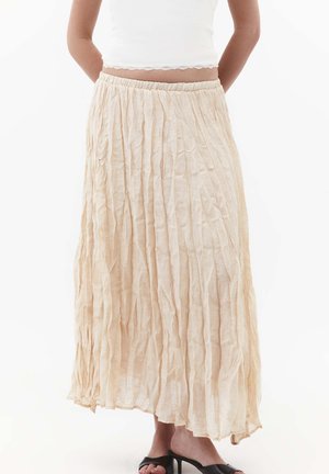 Donna che indossa una gonna lunga plissettata beige, un top bianco corto e sandali neri con tacco e punta aperta, in piedi con le mani dietro la schiena.