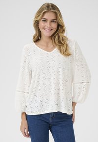 Blouse blanche avec un décolleté en V, ornée de motifs à œillets et de manches bouffantes avec des détails délicats. Associée à un jean bleu.