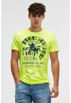 Junger Mann trägt ein leuchtend gelbes T-Shirt mit strandbezogenem Text und Palmenmotiv, kombiniert mit hellblauen Jeans, steht mit den Händen in den Taschen.
