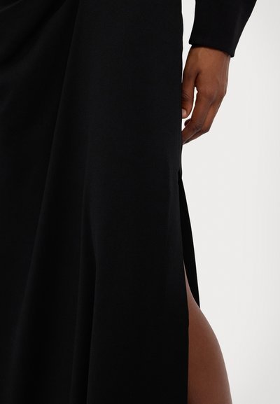 Schwarzes Kleid mit einem seitlichen Schlitz, fließendem Stoff mit einer glatten Textur. Die Hand, die sich nahe dem Schlitz positioniert, verleiht dem Design Details.