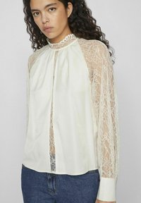 Blusa de encaje con cuello alto, mangas largas de encaje transparente y abertura en la parte delantera. La tela es de un crema claro con intrincados detalles de encaje.