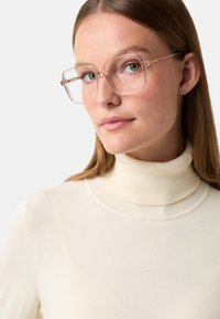 Žena s rovnými světle hnědými vlasy, která nosí průhledné oversize čtvercové brýle a krémový rolák, na jednobarevném pozadí.