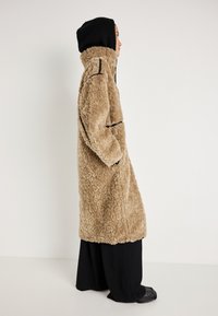 Beidzētā krēma krāsas awedzīgā mēteli ar strukturētu ārpusi, melnām akcentiem un kapuci. Mētelis ir līdz ceļiem un valkāts virs platām melnām biksēm.