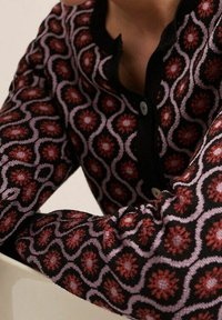 Cárdigan de punto con una base negra y un patrón floral en rosa y rojo, cuello redondo y cierre de botones. Mangas ajustadas y texturizadas.