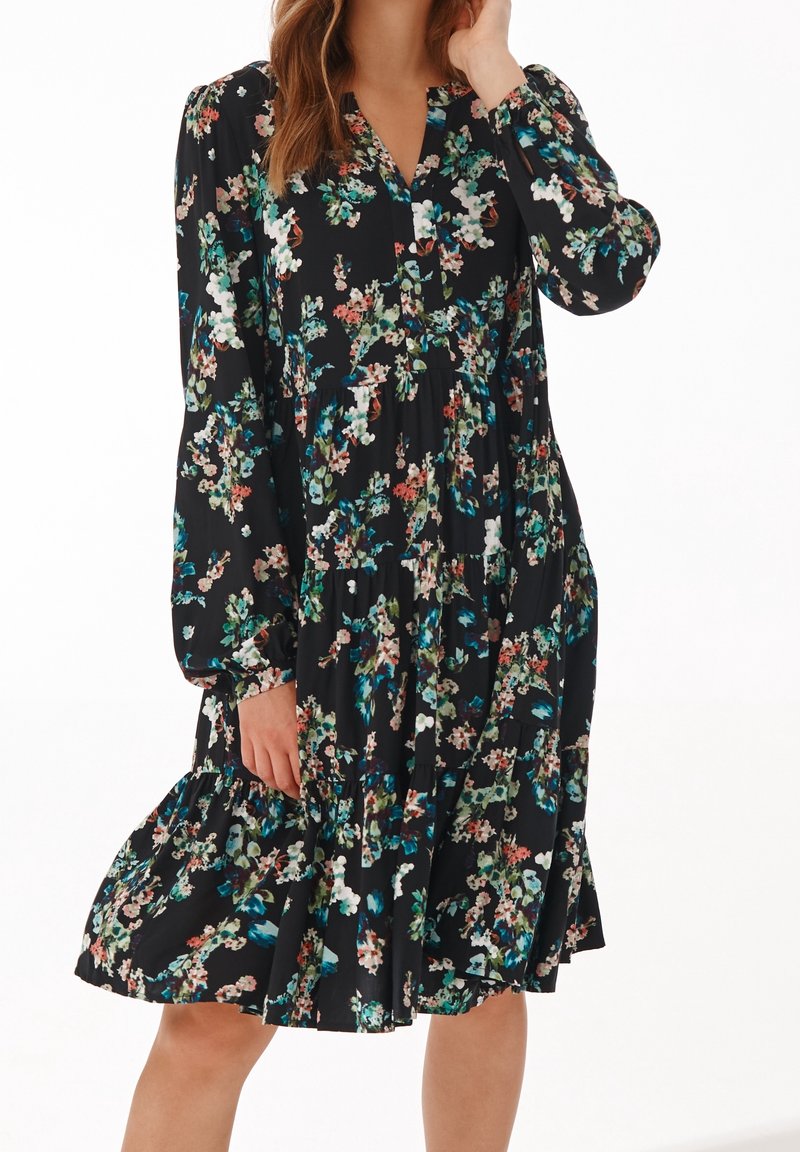 Femme portant une robe noire à fleurs, longue aux genoux, manches longues avec une jupe ample à volants et un col en V.