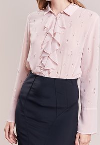 Helles rosa Bluse mit einem gerüschten Vorderteil, aus transparenter Stoff gefertigt. Verfügt über kleine metallische Punkte und lange Ärmel. Kombiniert mit einem enganliegenden schwarzen Rock.