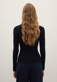 Pull en maille côtelée bleu marine avec manches longues, silhouette ajustée et encolure arrondie. Fond de couleur claire unie. Cheveux ondulés.