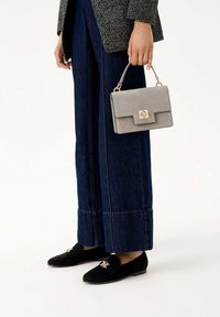 Borsa in suede grigia con hardware dorato, forma rettangolare e chiusura a patta, tenuta in mano con jeans larghi blu scuro e mocassini neri.