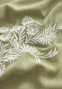 Tissu vert clair avec broderie florale blanche détaillée montrant feuilles et fleurs en gros plan.