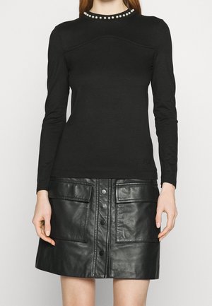 Vrouw met een zwart shirt met lange mouwen en parelbijzondere halslijn en een zwarte leren rok met knopen aan de voorkant en grote voorzakken.