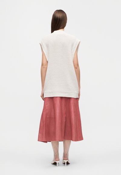 Haut sans manches tricoté beige avec bords côtelés, associé à une longue jupe en cuir rose et des collants en dentelle blanche, affichant un look en couches.