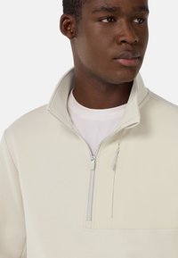 Pullover in pile con zip di colore beige chiaro, colletto alto, texture liscia, tasca decorativa sul petto e dettagli in hardware argentato.