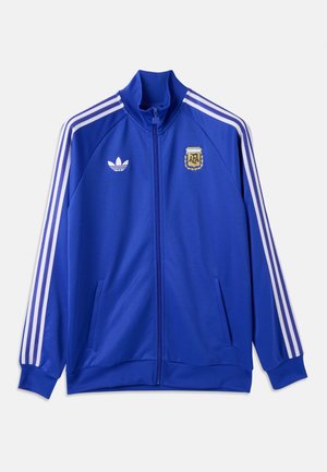 Jachetă sport albastră cu fermoar complet, cu dungi albe Adidas pe mâneci, logo-ul Adidas pe partea stângă a pieptului și emblema echipei de fotbal Argentina pe partea dreaptă a pieptului.