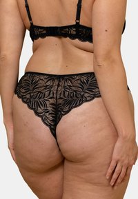 Ensemble de lingerie en dentelle noire avec des motifs floraux raffinés, une texture transparente et des bords festonnés, mettant en avant un style tanga.