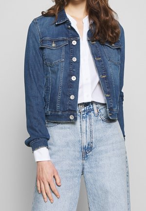 Kvinde iført en mørkeblå denimjakke over en hvid skjorte kombineret med høj taljede, lyseblå denimjeans, med hånden hvilende ved siden af sig.