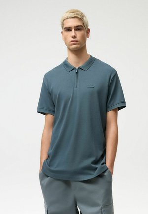 Młody mężczyzna z krótkimi blond włosami, ubrany w turkusową koszulkę polo na zamek i szare spodnie, stoi z rękami w kieszeniach na jednolitym tle.