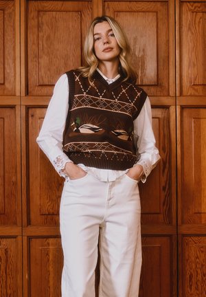 Blond naine seisab käed taskutes, kandes valget pluusi ja pükse, pruunist lõngast vestiga, millel tikitud pardid, taustal puidust paneel.