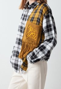 Chemise à manches longues à carreaux en tissu noir, blanc et jaune ; dotée d'un patch brodé moutarde avec des détails en franges.