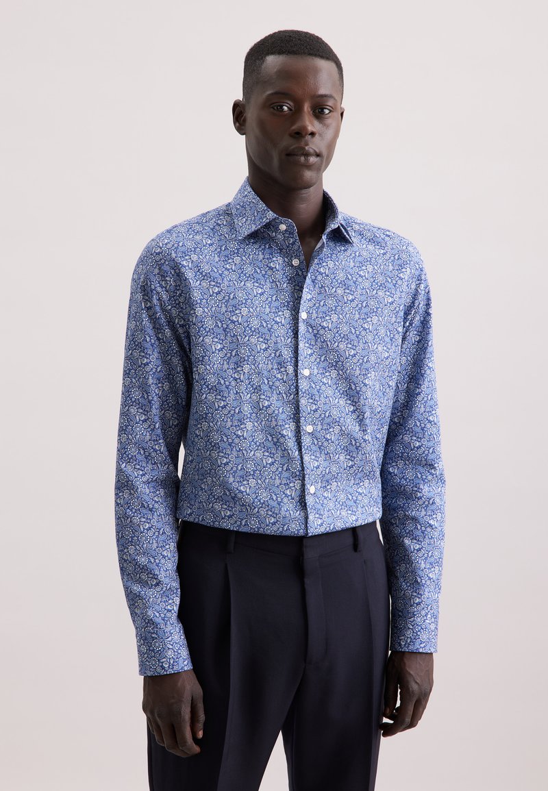 Camicia a maniche lunghe con motivo floreale blu e colletto button-down, realizzata in tessuto leggero, abbinata a pantaloni sartoriali scuri.