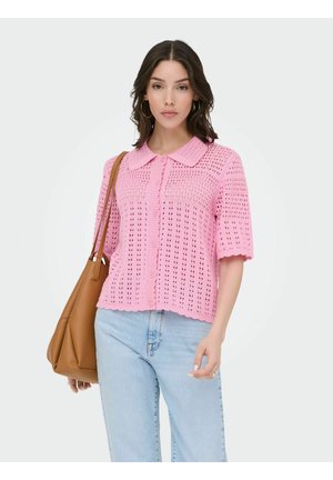 Nő rózsaszín horgolt rövid ujjú felsőben, világoskék farmerral, nagy karika fülbevalókkal, és barna bőr válltáskával a vállán.