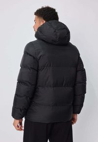 Veste matelassée noire avec capuche, design matelassé et matériau isolant. Vue de dos montrant des épaules arrondies et des poignets élastiques.