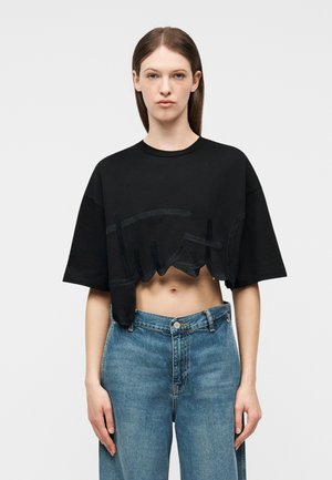 Γυναίκα με ίσια καστανά μαλλιά φοράει μια μαύρη cropped oversized μπλούζα με κεντημένα γράμματα και τζιν ψηλόμεσο σε μπλε απόχρωση, στέκεται μπροστά σε λευκό φόντο.