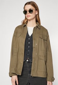 Olive green, oversized jacket with a button front and side pockets. Worn over a black vest, paired with round sunglasses.