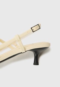Sandal tinggi berhak beige dengan tali, memiliki dasar hitam. Dihiasi dengan tali tipis dan gesper kotak kecil dengan perforasi untuk penyesuaian ukuran.