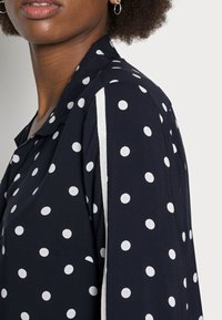 Camicia navy a pois con pois bianchi, colletto classico e un dettaglio laterale con una striscia a contrasto. Tessuto leggero.