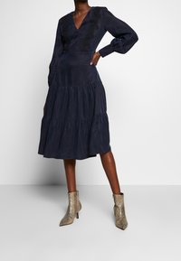 Mörkblå klänning med v-ringning och långa ärmar, med broderade detaljer och en flerlagerskjol, i kombination med beige mönstrade ankelstövlar.