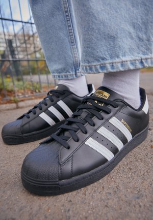 Sort Adidas Superstar sneakers med hvide striber, båret med hvide sokker og lyseblå jeans, stående på en fortov udendørs.