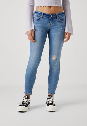 Femme portant un jean skinny bleu clair déchiré, un crop top blanc à manches longues et des baskets noires et blanches, debout sur un fond uni.
