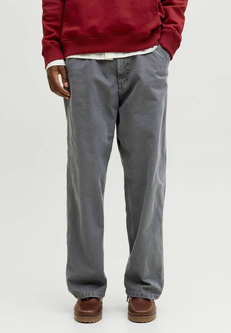 Pantaloni di cotone grigi con una vestibilità ampia e taglio dritto, dotati di tasche laterali. Indossati con una felpa rossa e scarpe marroni. Tessuto testurizzato.