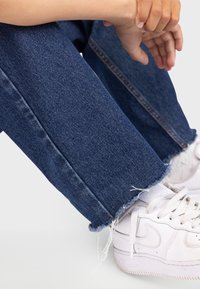 Jean en denim bleu avec un ourlet effiloché, associé à des baskets blanches. Le denim a une texture lisse et un design à coupe droite classique.