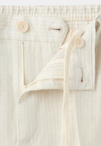 Ceinture de pantalon beige avec élastique, fines rayures verticales, fermeture éclair partiellement ouverte et deux boutons beiges sur les boutonnières.