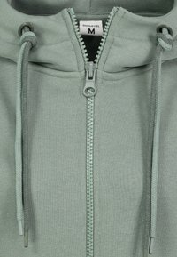 Hellgrüner Reißverschluss-Hoodie aus weichem Stoff. Verfügt über einen vorderen Reißverschluss, verstellbare Kordeln und ein Etikett, das die Größe M angibt.