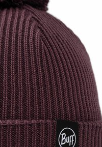 Purple knitted beanie with a ribbed texture, topped with a fluffy pom-pom. Features a black logo patch with white lettering.