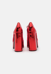 Tacones altos de charol rojo con un tacón cuadrado grueso, punta afilada y acentos de malla en los laterales. Superficie lisa y brillante.