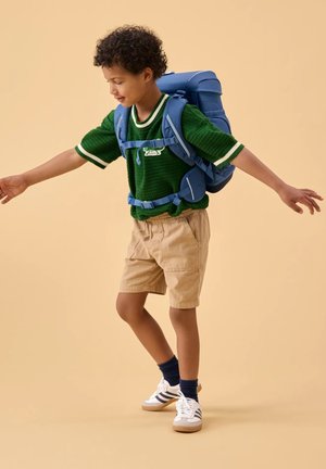 Junge mit lockigem Haar, der einen grünen Pullover, beige Shorts, weiße Turnschuhe und einen blauen Rucksack trägt und mit ausgestreckten Armen vor einem beigen Hintergrund balanciert.