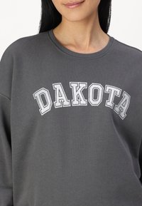 Sudadera gris con cuello redondo que presenta la palabra "DAKOTA" en letras gruesas blancas y grises en el pecho, hecha de un tejido suave.