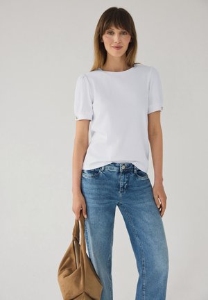 Kvinde iført hvid bluse med korte ærmer og blå jeans, der holder en tan ruskindshåndtaske ved siden af sig mod en ensfarvet baggrund.