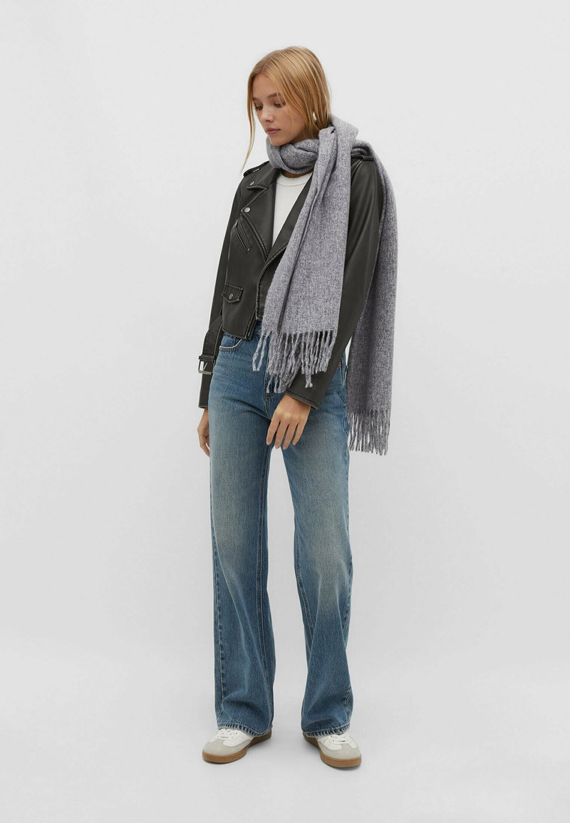 Écharpe grise avec des franges, veste en cuir de motard, top blanc, jeans bleus évasés et baskets blanches. Accent sur les textures et les couleurs.