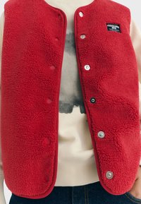 Gilet en polaire rouge avec des boutons-pression, présentant une texture pelucheuse et un empiècement contrasté. Porté par-dessus un sweat-shirt de couleur claire avec un motif graphique.