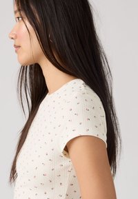 Νεαρή γυναίκα με μακριά σκούρα μαλλιά, φοράει λευκό μπλουζάκι με κοντά μανίκια και μικρά ροζ ανθισμένα σχέδια, εμφανίζεται σε προφίλ πάνω σε απλό φόντο.